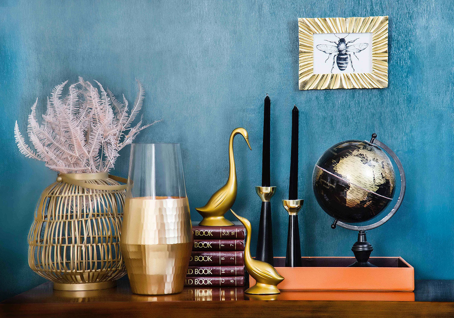 shelf with burgundy books gold bird statues black candles and globe on top with blue wall behind it and illustration of bee hanging on wall